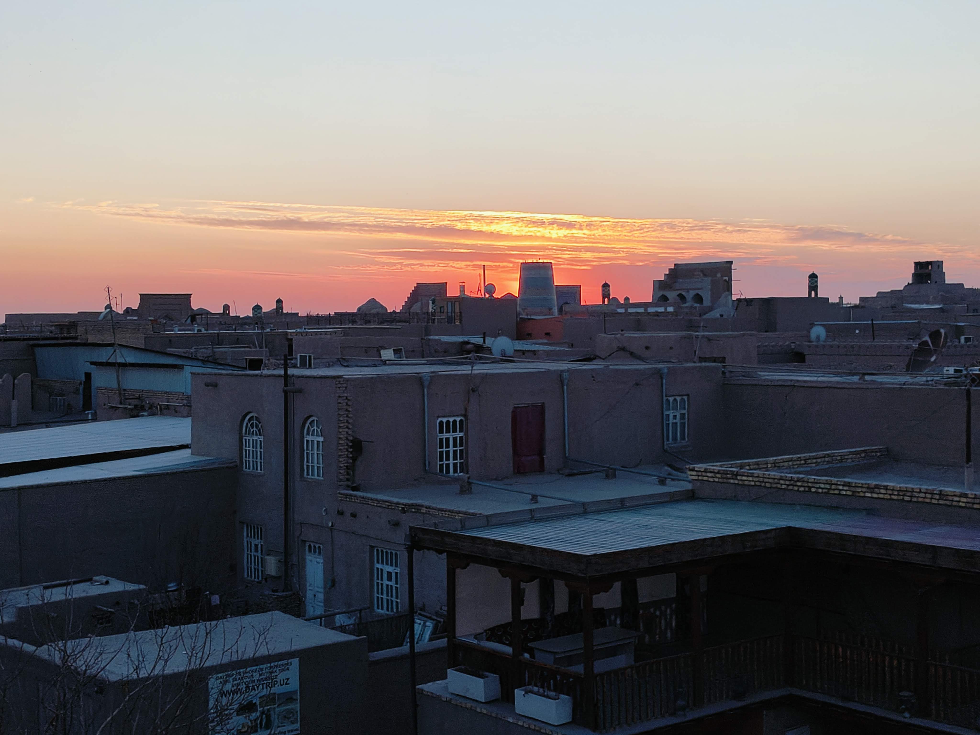 Dramatic sunset over Khiva's ancient skyline from city walls - silhouettes of historic minarets and domes against vibrant orange sky in Uzbekistan's oldest Silk Road city
